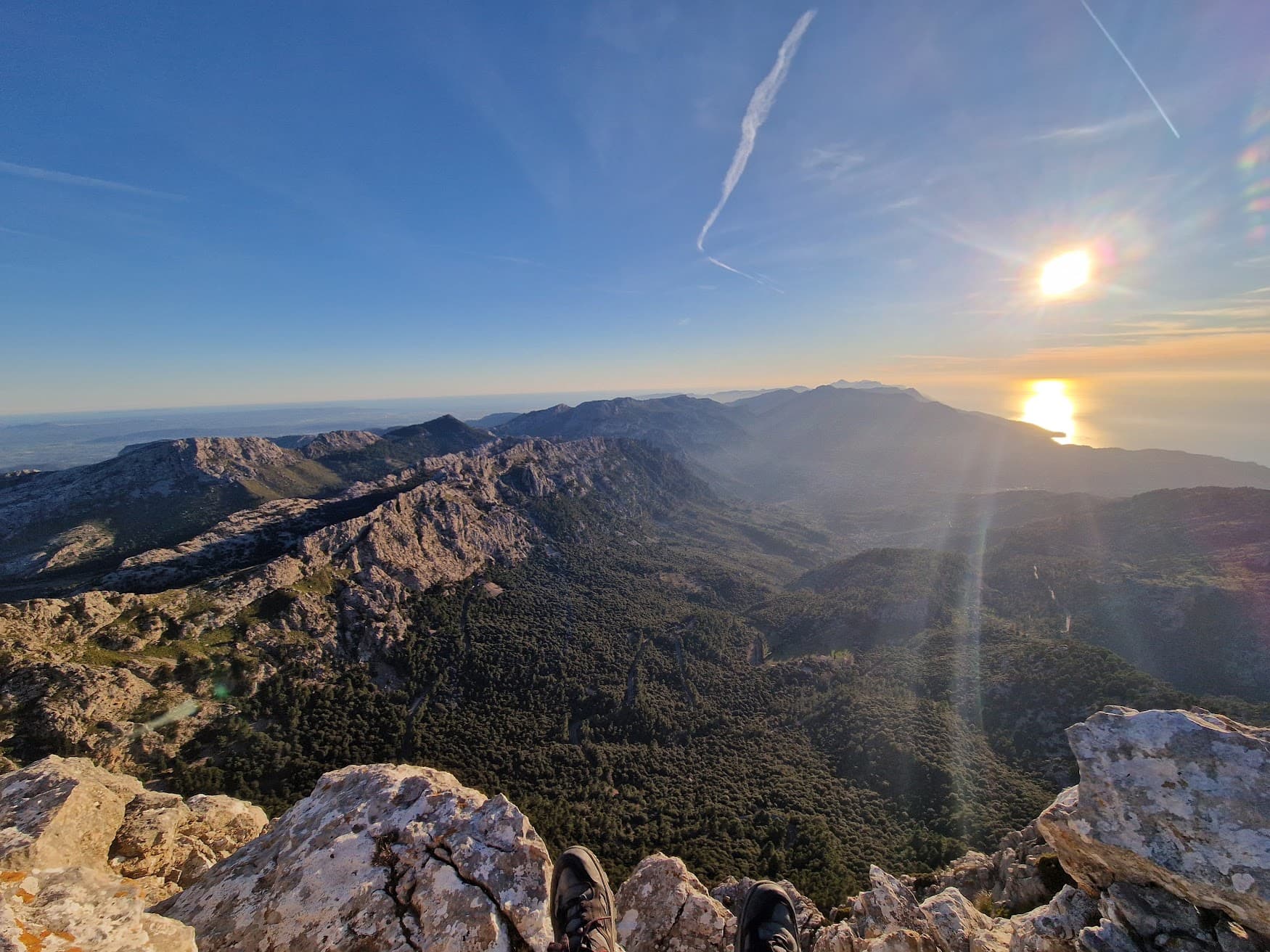 Hiking boots on a rocky peak overlooking a vast mountain range and sunlit coastal horizon.