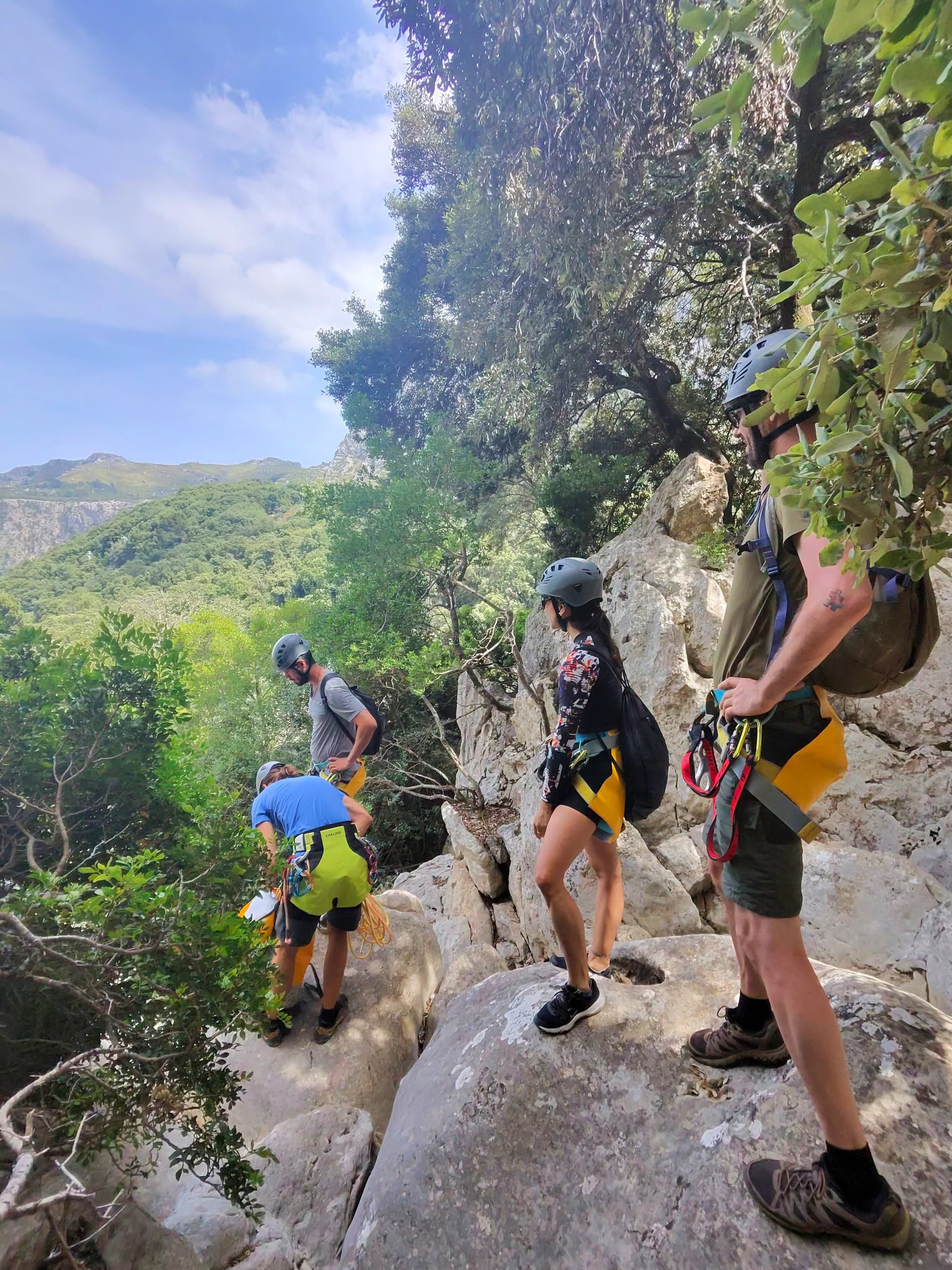 Four climbers in helmets and harnesses stand on a rocky ledge overlooking a lush valley.