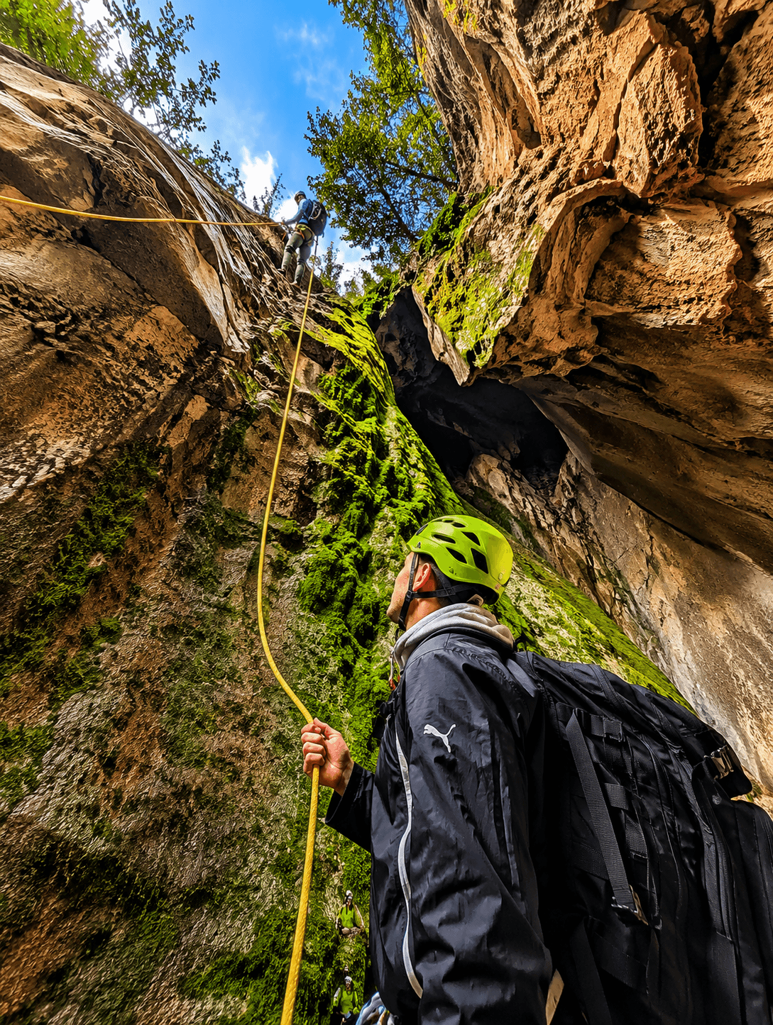 Climber in a green helmet holds a rope while another rappels down a mossy rock wall.