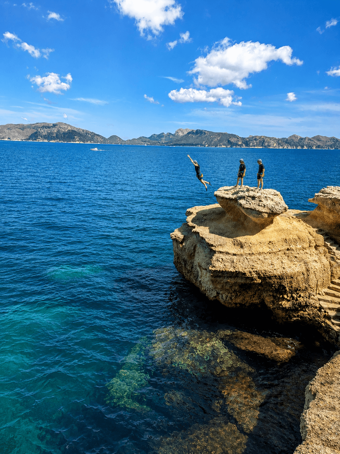 Person jumping from a rocky cliff into the blue sea with two onlookers watching.