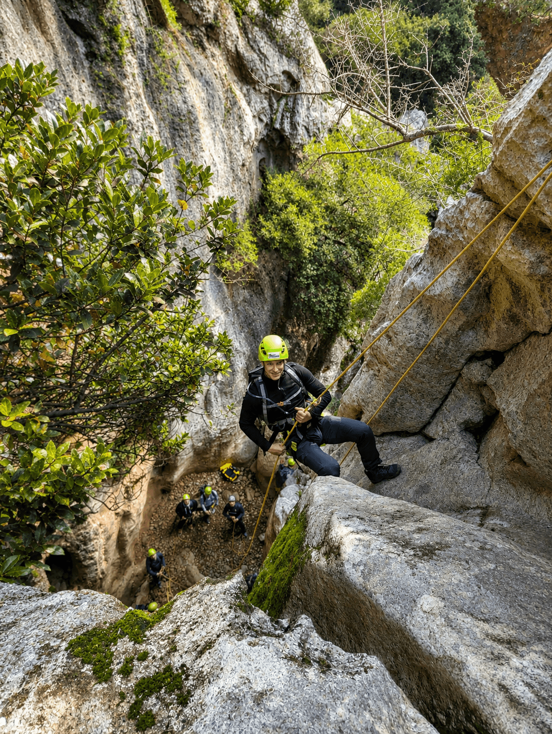 A smiling man in a lime green helmet rappels down a steep, rocky cliff face.