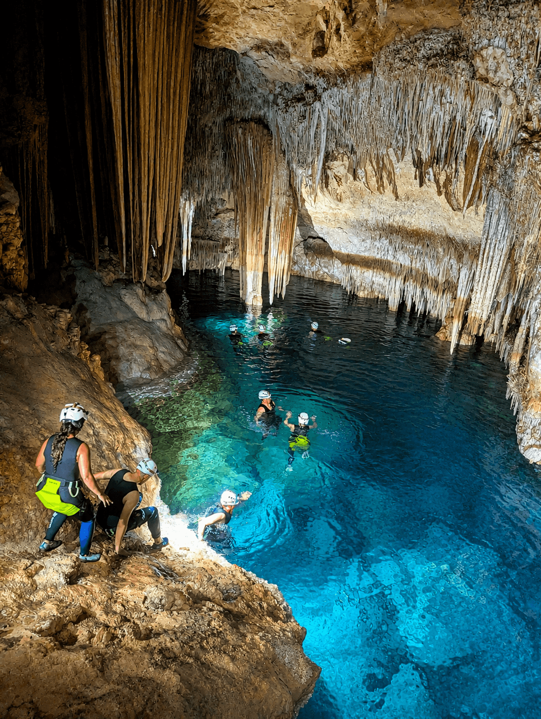 Adventurers swim in clear blue water within a limestone cave featuring dramatic stalactite formations.