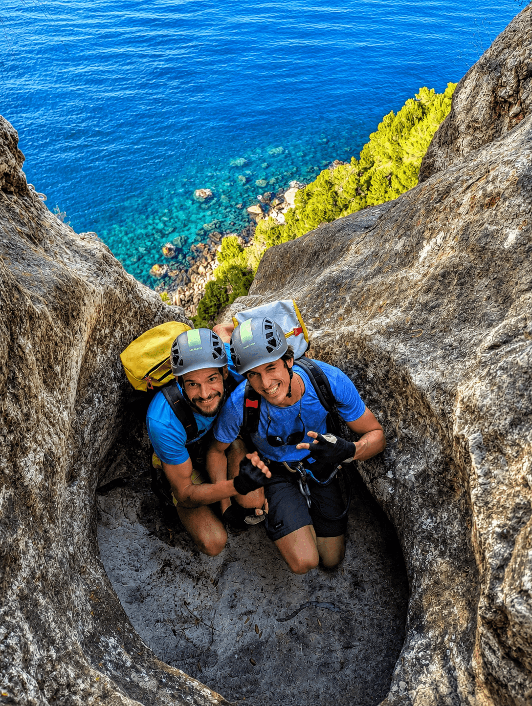 Two smiling climbers in gear inside a rocky crevice overlooking a bright blue sea.