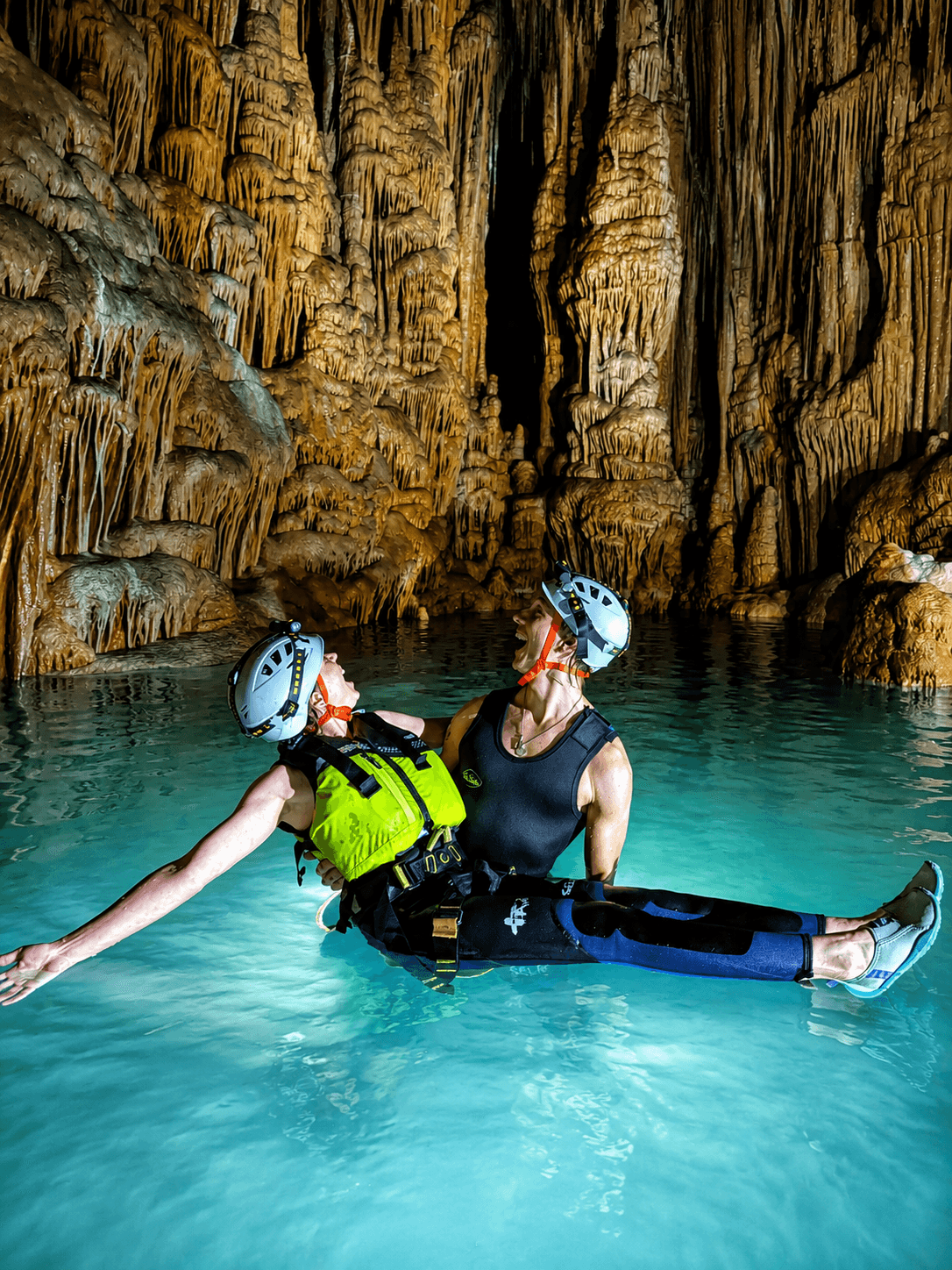 Two explorers laughing in turquoise water inside a cave with dramatic stalactite and stalagmite formations.