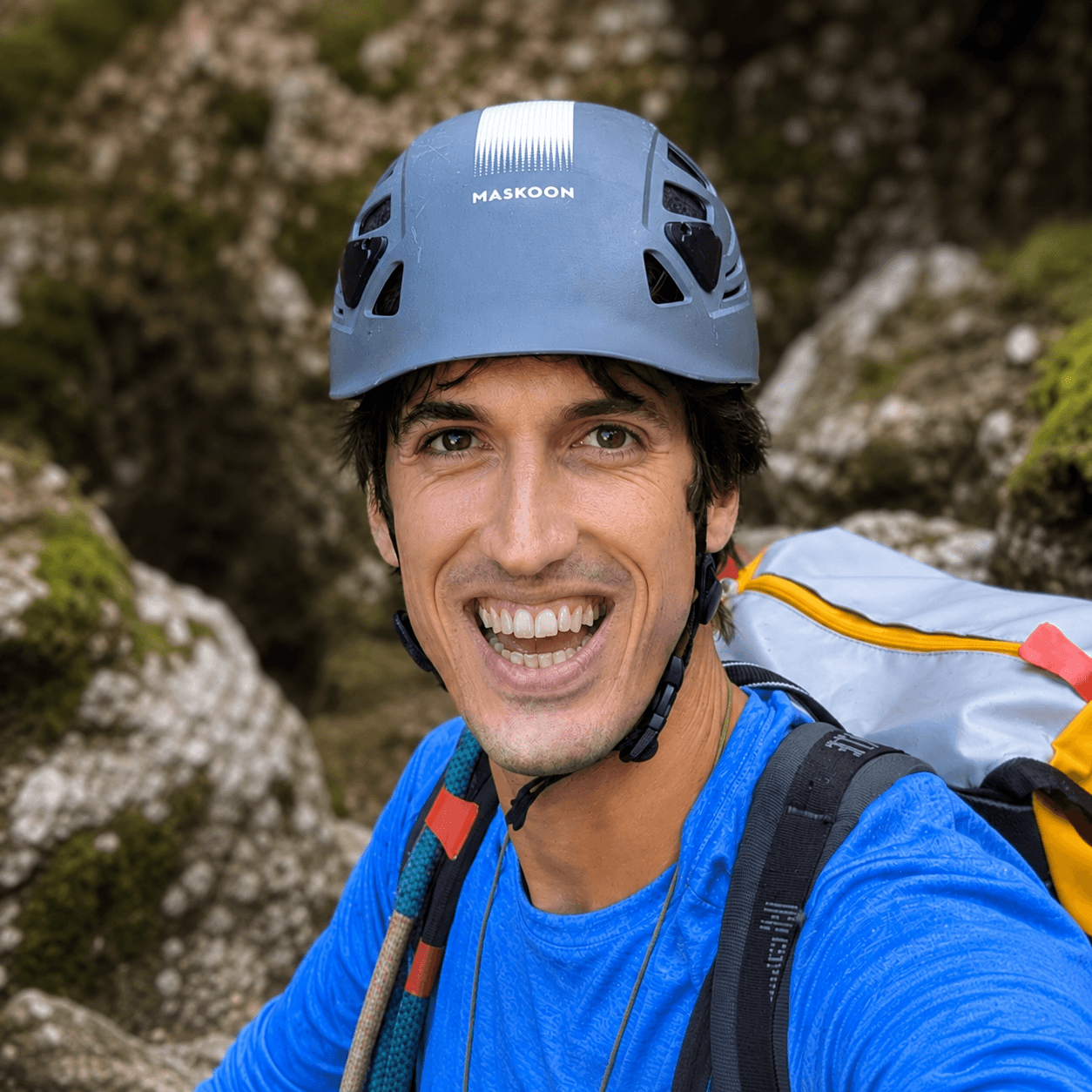 Smiling man wearing a blue climbing helmet and backpack in a rugged, rocky landscape.